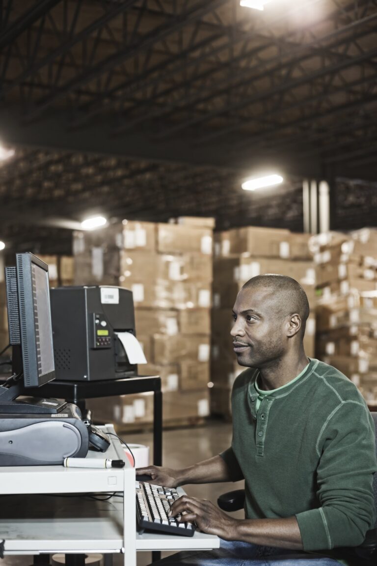 African American male warehouse worker working on a desktop computer in a large distribution